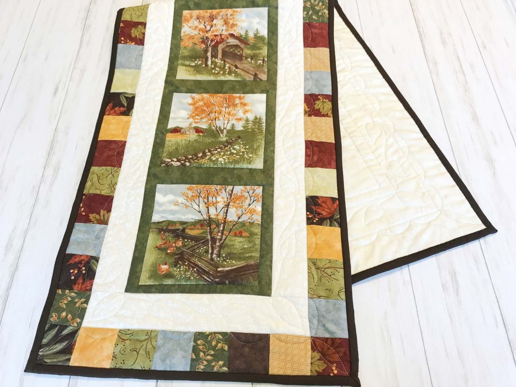 Quilted Table Runner folded over with three scenes of a covered bridge, a church in a fields and an old fence with an outer border.